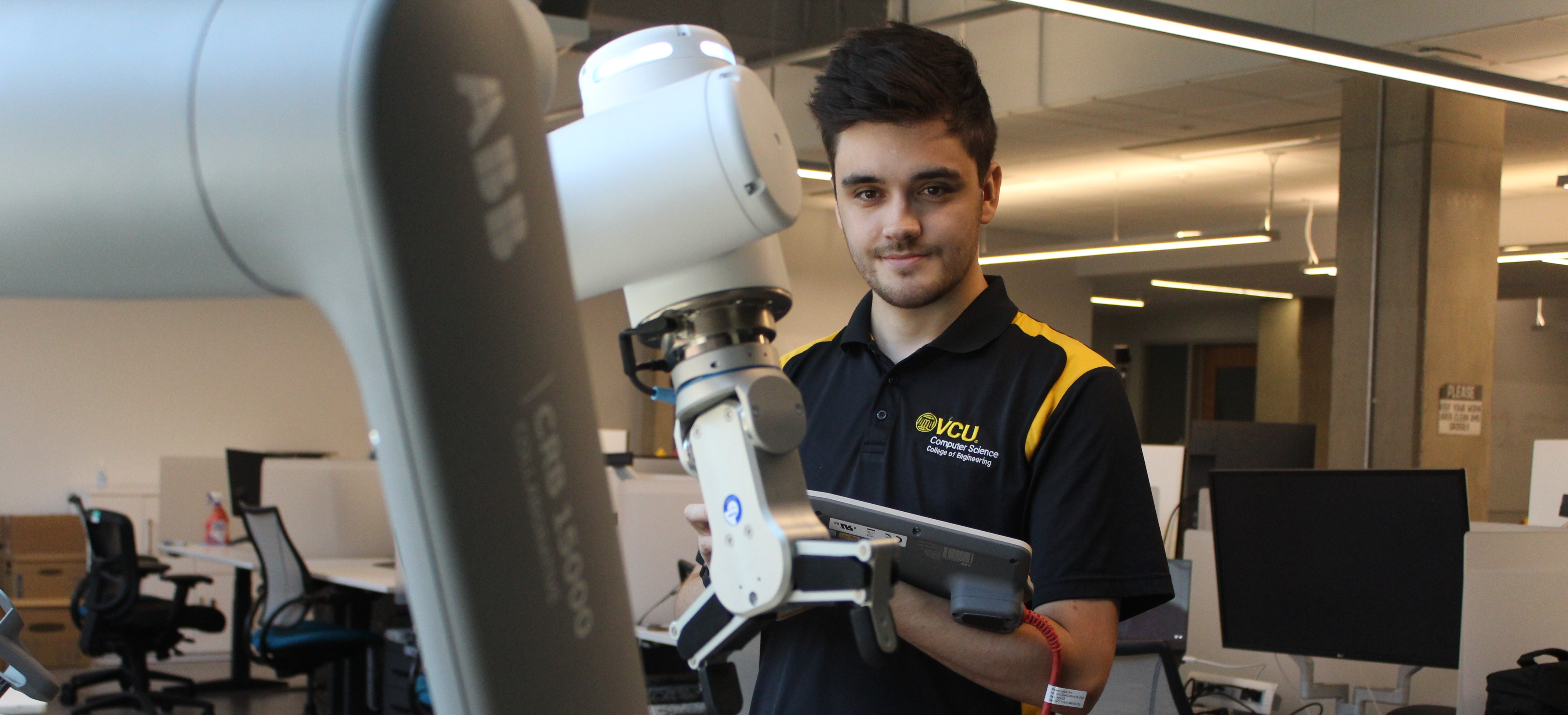 A student using a VCU shirt is manipulating a teaching pendant to control a one-armed collaborative robot.