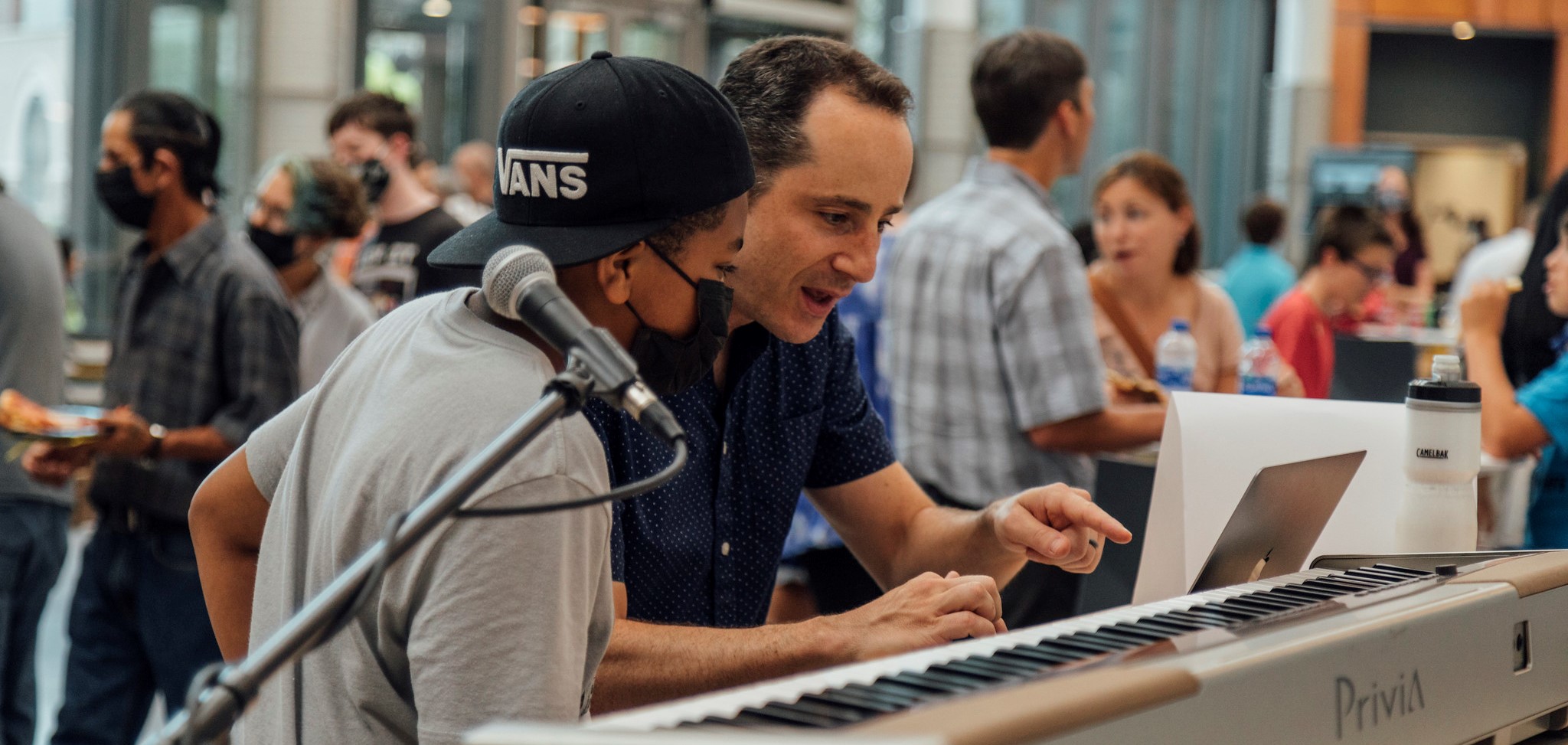 Professor and student looking at an electronic keyboard. Professor is pointing to the keys of the keyboard while the student watches his movements.